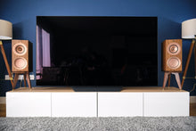 Load image into Gallery viewer, Large flat-screen TV on a white stand with wooden KEF R3 speakers on special height speaker stands by Audio Chic against a blue wall.