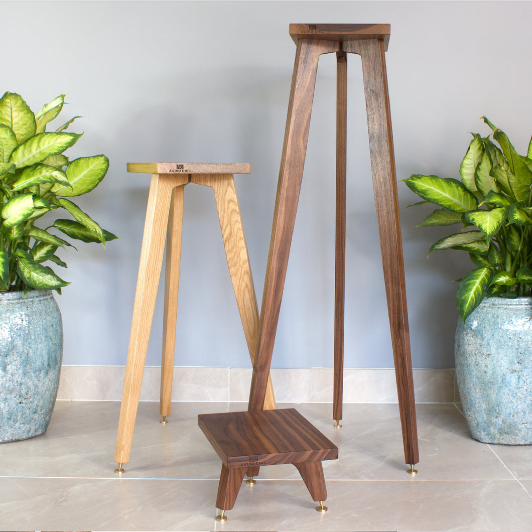A hardwood speaker stand with a tripod design, displayed alongside two potted plants. The stand is solid and has a natural wood finish.