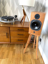 Load image into Gallery viewer, Wooden speaker on a speaker stand by audio chic next to a wooden cabinet with a geometric wall pattern.