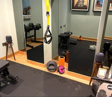 Load image into Gallery viewer, Apple homepod speakers on a walnut speaker stands by audio chic in a Gym room with exercise equipment including a bench, weights, and mirrors.