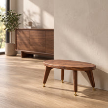 Load image into Gallery viewer, A mid-century modern oval wooden speaker stand by Audio Chic with tapered legs and brass spikes and discs, positioned in a minimalist room with light wood floors and a textured beige wall.