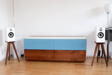 Load image into Gallery viewer, B&amp;W 607, 607 S2 and 607 S3 speaker stands in walnut in a living room. Modern room setup with a blue and wood cabinet, white speakers on stands, and a white wall.