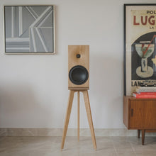 Load image into Gallery viewer, A wooden Buchart Audio E50-S speaker on an Audio Chic tripod speaker stand in a minimalist room setting with framed wall art and a wooden cabinet.