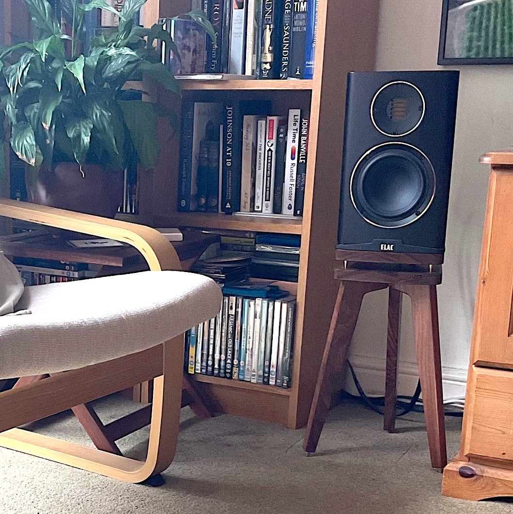 Elac Carina Speaker on a Walnut Audio Chic speaker stand in a room setting next to a bookcase and comfy chair