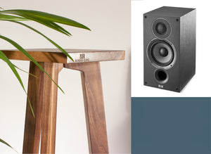 An Elac Debut B5.2 speaker with a wooden stand with a tripod base, with a plant in the background and Audio Chic brand logo on the front