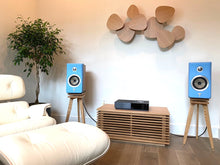 Load image into Gallery viewer, A modern home audio setup featuring blue Focal Kanta N1 speakers on wooden speaker stands by Audio Chic in a living room.