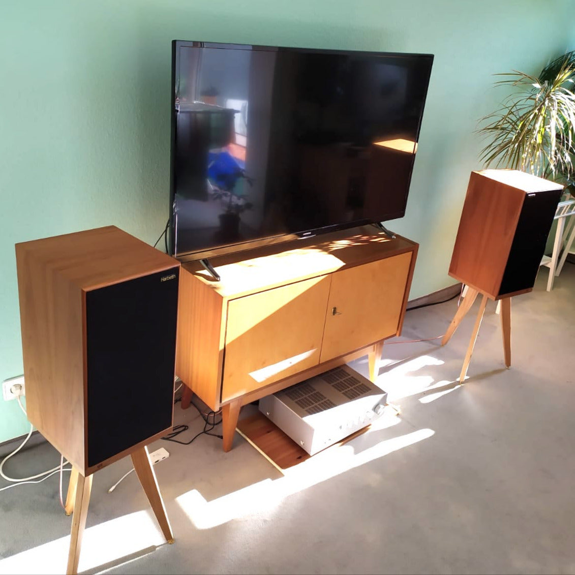Living room with a large TV on a wooden stand, flanked by Harbeth C7ES3 speakers on Cherry Speaker stands from Audio Chic, against a light green wall.