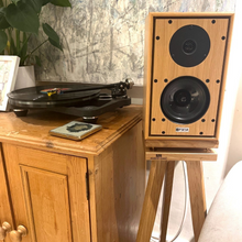Load image into Gallery viewer, Harbeth-P3ESR-Speaker-Oak-Heron-speaker-stand-by-audio-chic. Wooden speaker next to a turntable on a wooden cabinet with a marble wall in the background.