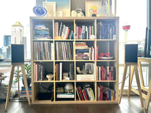 Load image into Gallery viewer, Harbeth-P3ESR-heron-speaker-stands-in-maple-by-audio-chic and a Bookshelf filled with books and decorative items in a room with large windows.