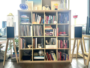 Harbeth-P3ESR-heron-speaker-stands-in-maple-by-audio-chic and a Bookshelf filled with books and decorative items in a room with large windows.