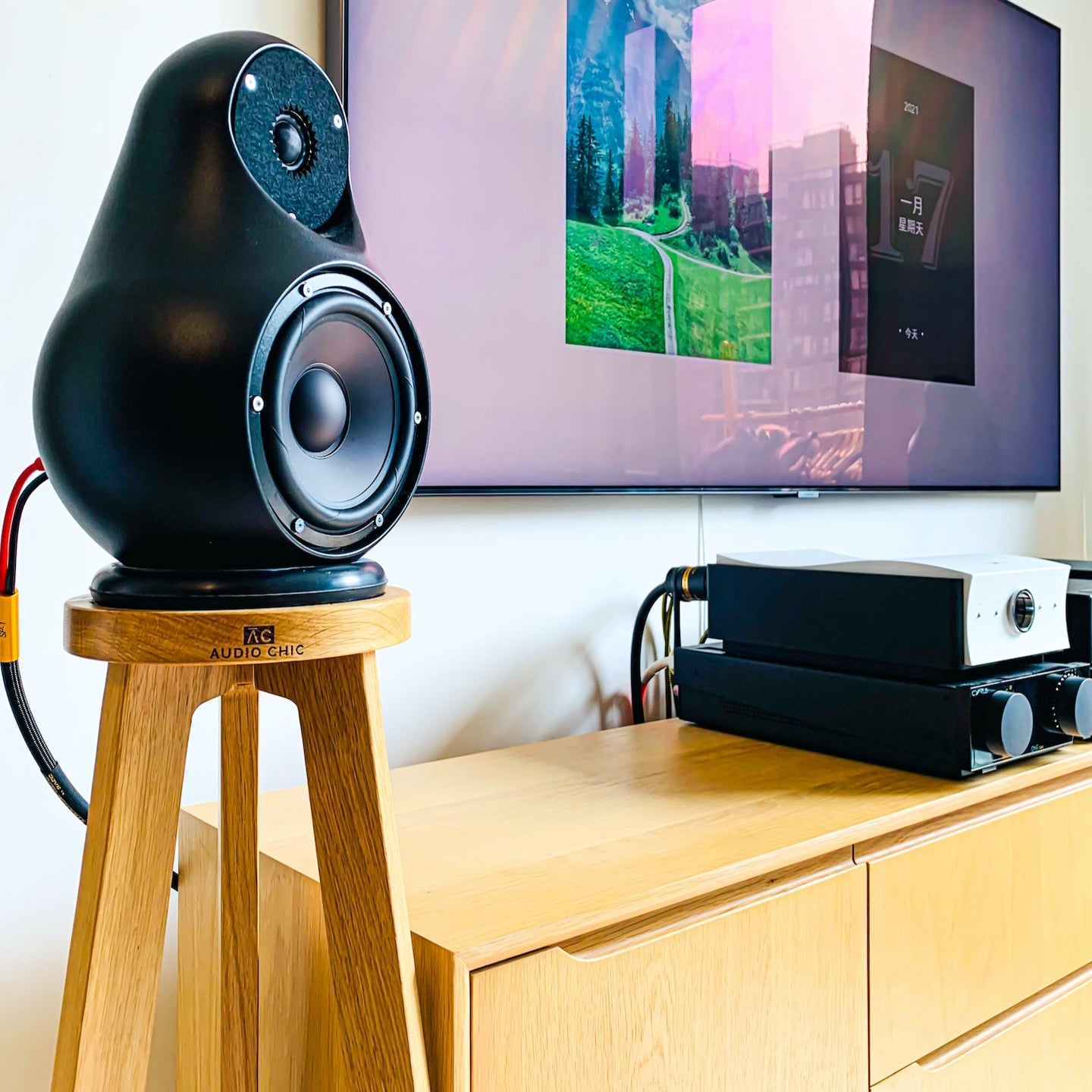 Black jern speaker on a shaped wooden speaker stand by audio chic with a television in the background