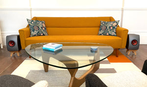 KEF-LS50W-II-Kite-speaker-stands-from-audio-chic in a Modern living room with an orange sofa, glass coffee table, and decorative items.