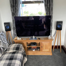 Load image into Gallery viewer, A pair of KEF LS50W speaker stands in a room, with one stand next to each speaker on a wooden TV stand. The stands are made of a light-colored wood and have a tripod design with an additional isolation plinth.