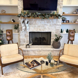 Christmas Living room with a stone fireplace, beige chairs, and a glass coffee table. With KEF R3 speakers on Walnut speaker stands by Audio Chic in the background.