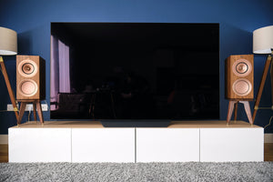 Large flat-screen TV on a white stand with wooden KEF R3 speakers on special height speaker stands by Audio Chic against a blue wall.
