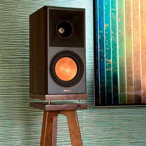 Klipsch-RP500M-II-Walnut-700-speaker-stand-by-audio-chic against a textured wall with a colourful abstract painting.