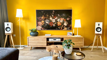 Load image into Gallery viewer, Living room with yellow walls, a TV, Martin-Logan-Motion-XT-B100-Oak-600-speaker-stand-by-audio-chic, and decorative items.
