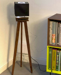 Naim-Audio-Mu-so-Qb-2nd-generation-Speaker_Stands-in-maple-by-audio-chic tripod stand against a white wall with a bookshelf in the background.
