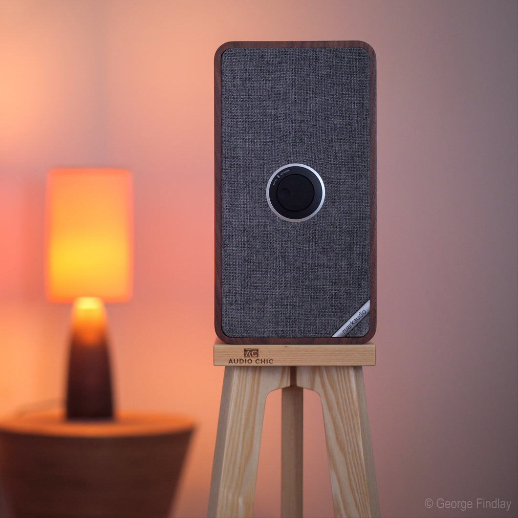 Ruark-Audio-MRX-Crane-speaker-stand-by-audio-chic-cropped with a blurred background featuring another speaker and a lamp.