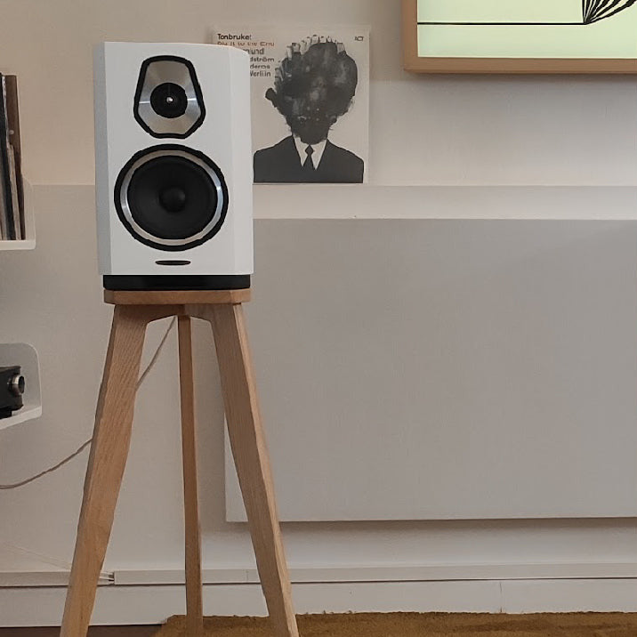 The image shows a shaped Sonus Faber Sonetto I speaker stand in oak finish from Audio Chic. One stand is shown in profile with the shaped speaker mounted on it, displaying the three-legged design and solid brass speaker spikes.