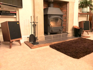 Wharfedale-Denton-85-drunken-snipe-in-walnut. In a room setting with a stove fire and rug. Speaker-stands-by-audio-chic