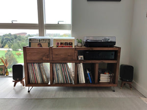 Q-acoustics-3010i-snipe-speaker-stands-in-walnut-by-audio-chic with a Wooden record player console with vinyl records and speakers in a room with large windows.