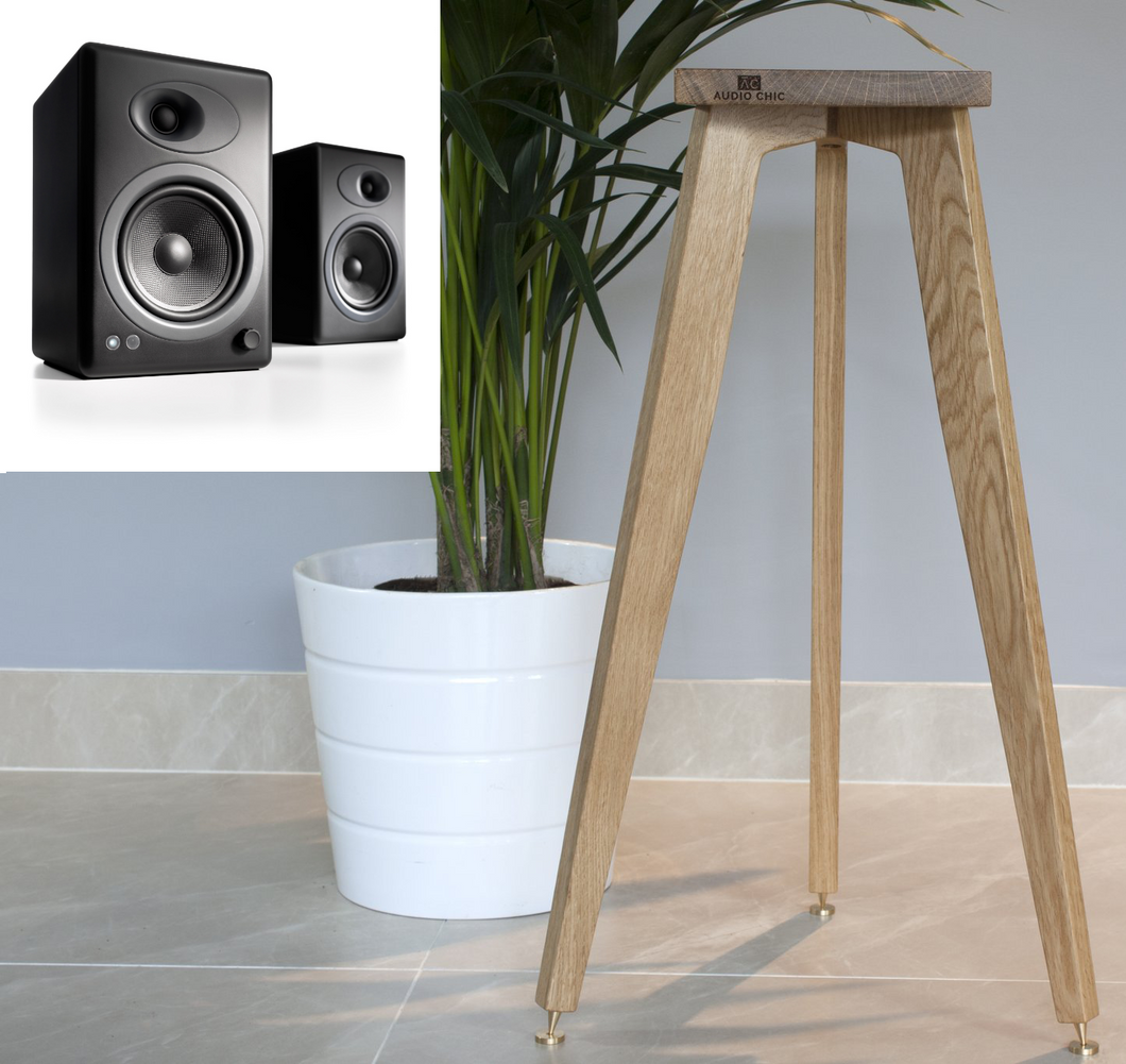 A pair of wooden speaker stands by Audio Chic with a single speaker placed on top, set against a home interior background. The stands have a natural wood finish and three legs each.