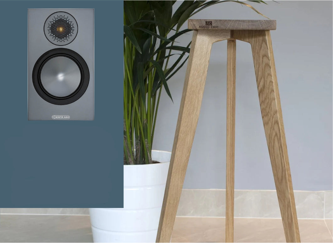 Monitor Audio Bronze 50 Speaker Stand in Oak wood with a speaker, positioned against a grey background with a plant beside it.