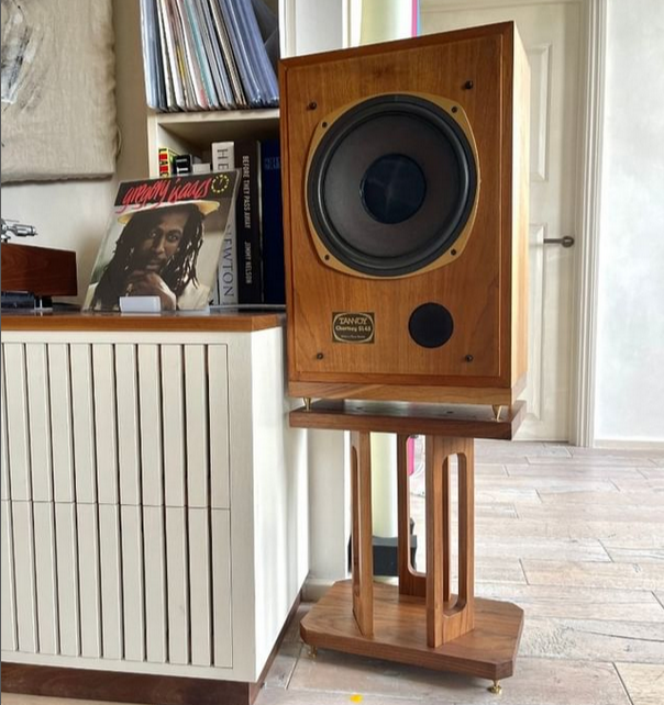 A wooden floor-standing speaker on top of an adjustable stand, placed next to a record player and books, with a door in the background.