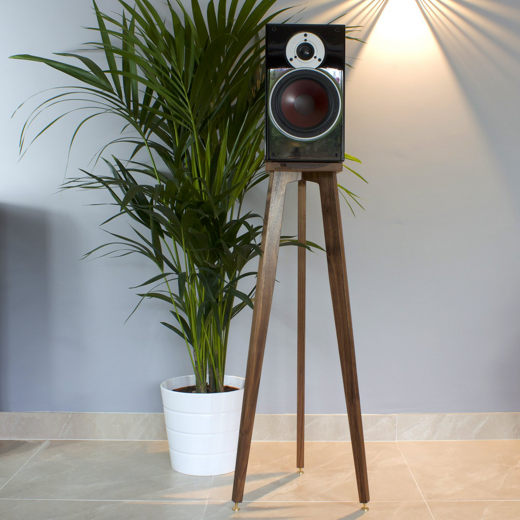 A pair of speaker stands made of walnut wood, with a speaker mounted on the top, placed next to a potted plant. The stands have a tripod design and are finished with a hard wax.
